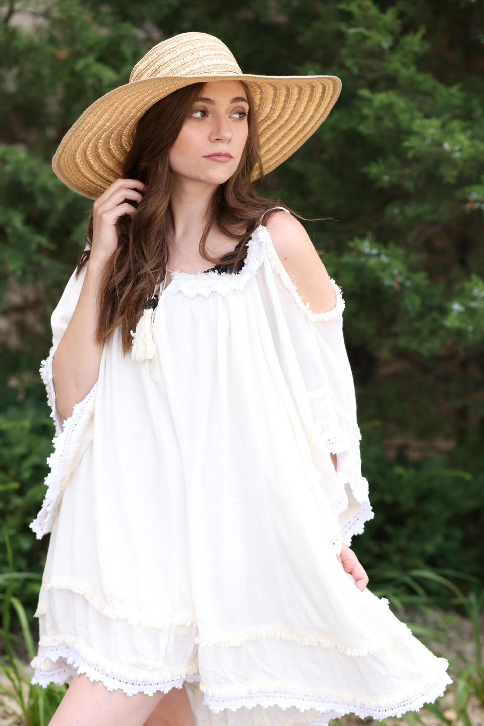 A portrait shot of a long haired brunette woman looking off to the side at a 3/4ths angle, she’s wearing a white dress with fringe and a sun hat. She’s holding the skirt of the dress in one hand, and her other hand is holding some of her hair. The backgrou